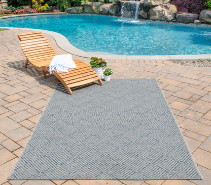 Blue geometric outdoor rug on tan stone-paver patio beside a wooden sun lounger by the pool, with towel and potted plant.