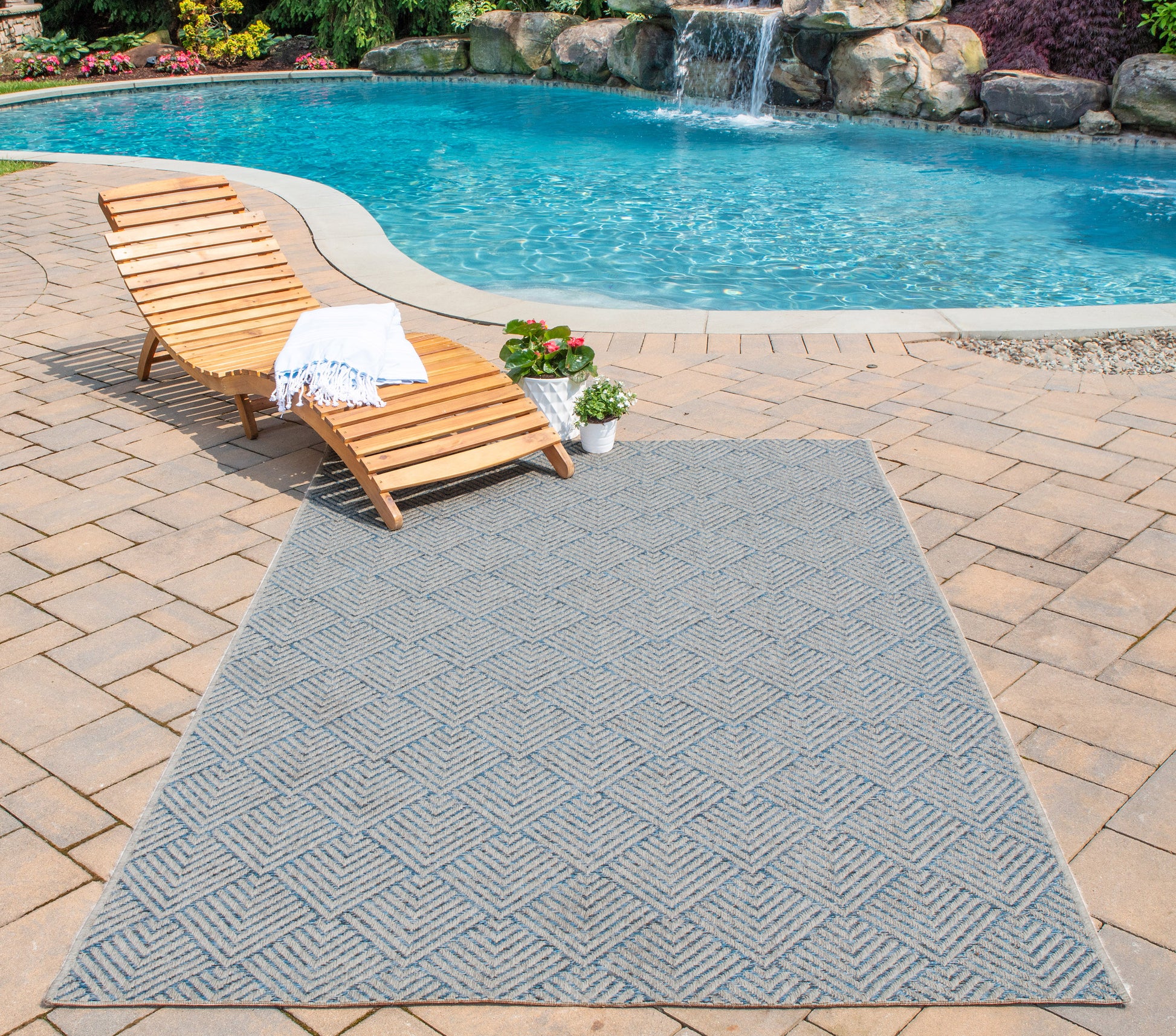 Blue-gray outdoor rug with a geometric diamond pattern on a pool deck beside a wooden chaise lounge.