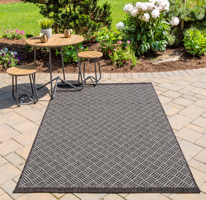 Outdoor charcoal-gray diamond-pattern rug on brick pavers with round wooden tables and stools; flowers in background.