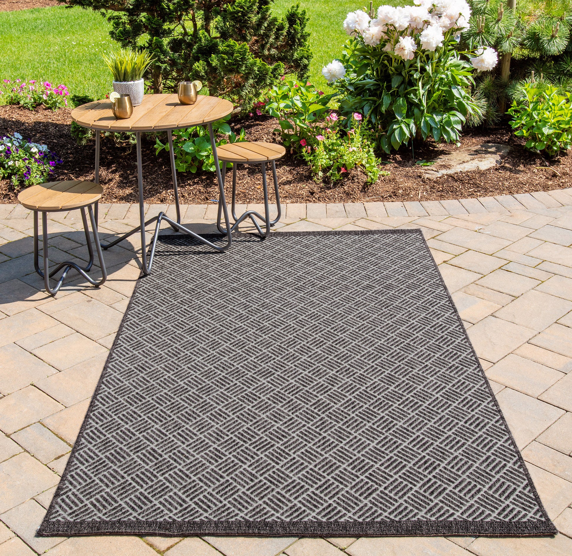 Outdoor gray diamond-pattern runner rug on a stone patio with round wood-topped tables and stools amid a lush garden.