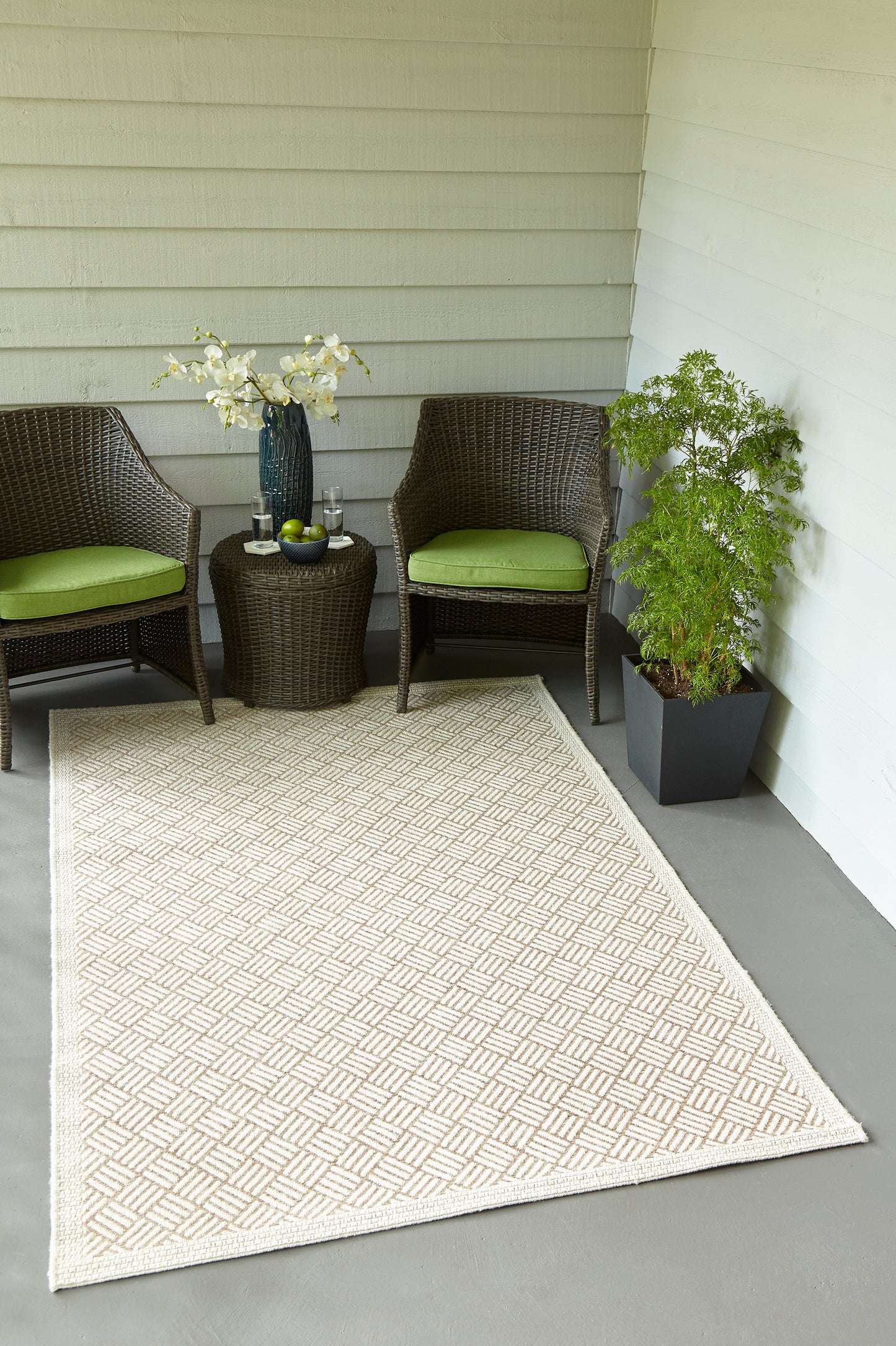 Outdoor wicker seating with lime cushions, round side table, vase of white flowers, beige geometric outdoor rug, potted plant.