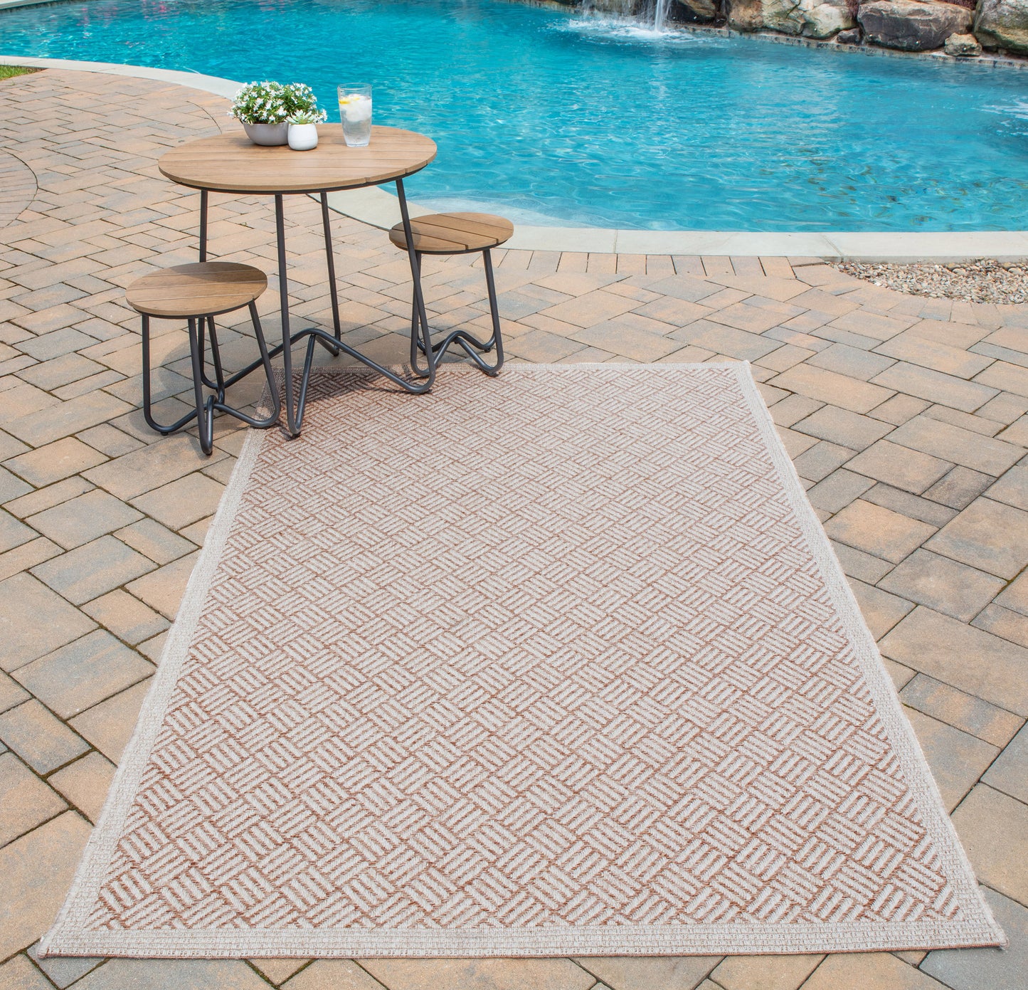 Outdoor tan geometric woven rug on brick-paver poolside patio with round wood-top table and two matching stools.