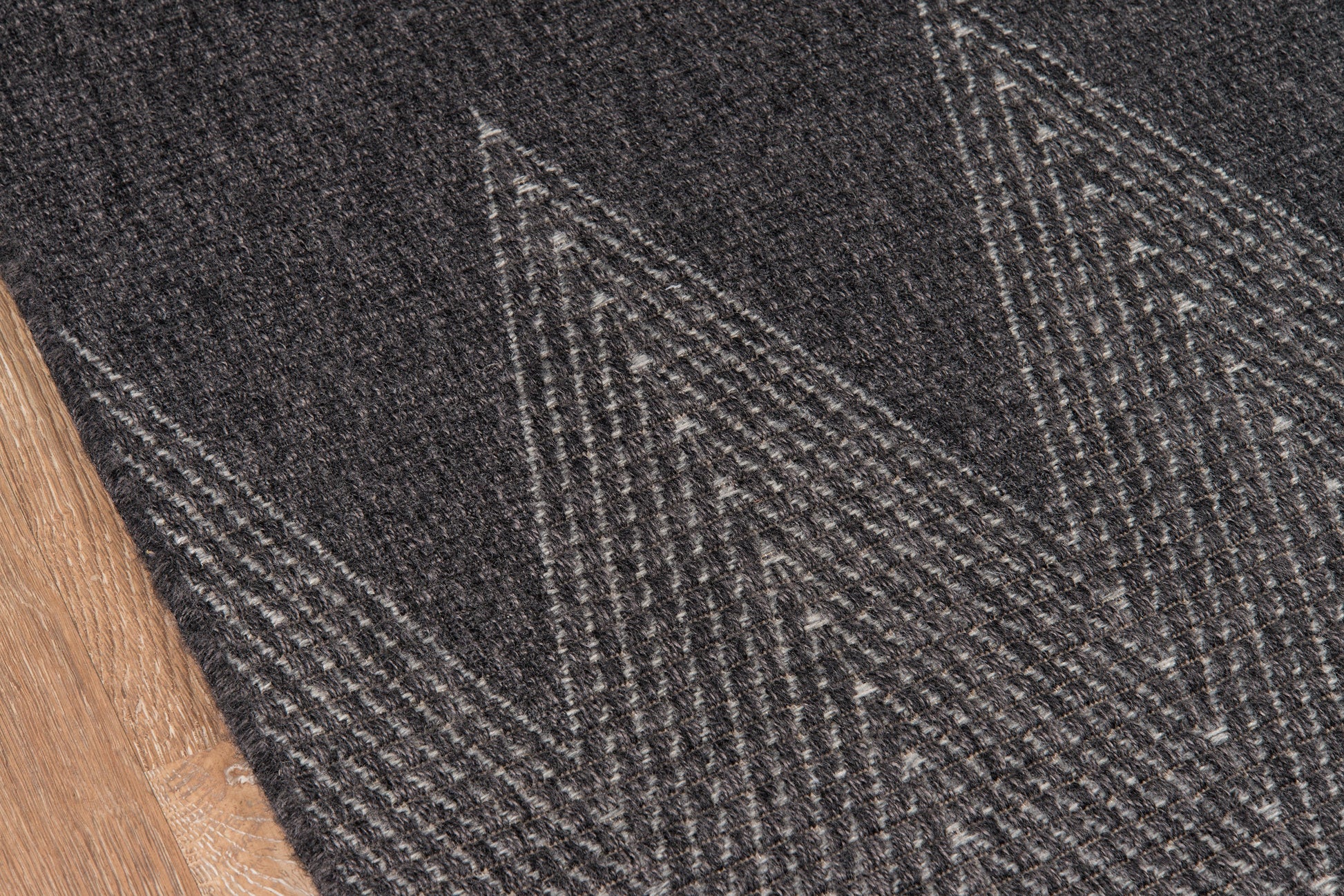 Dark charcoal woven rug with light gray chevron geometric pattern; wooden floor edge visible.