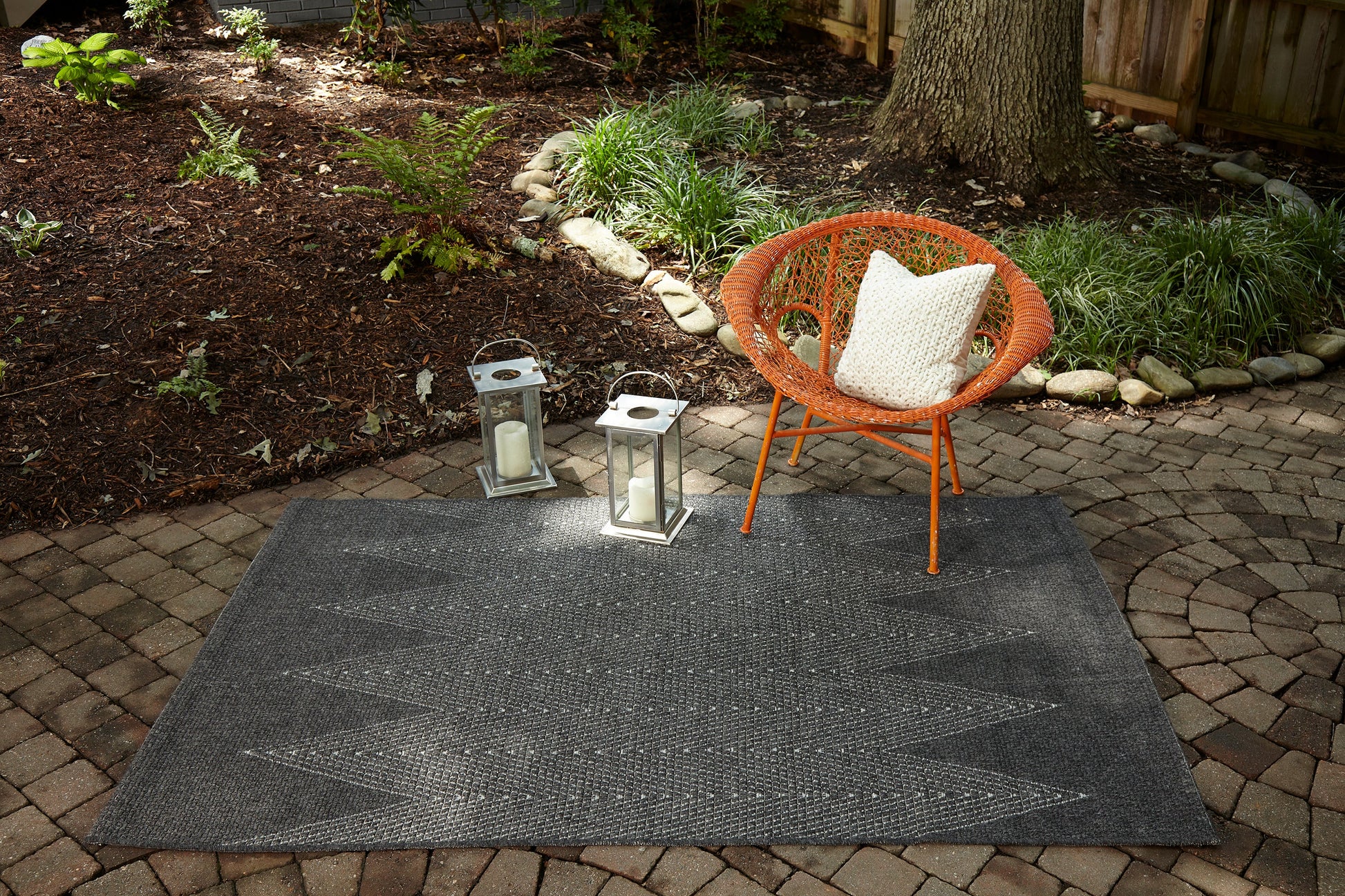 Outdoor patio with dark chevron rug, orange wicker chair with white pillow, and two metal lanterns on brick pavers.