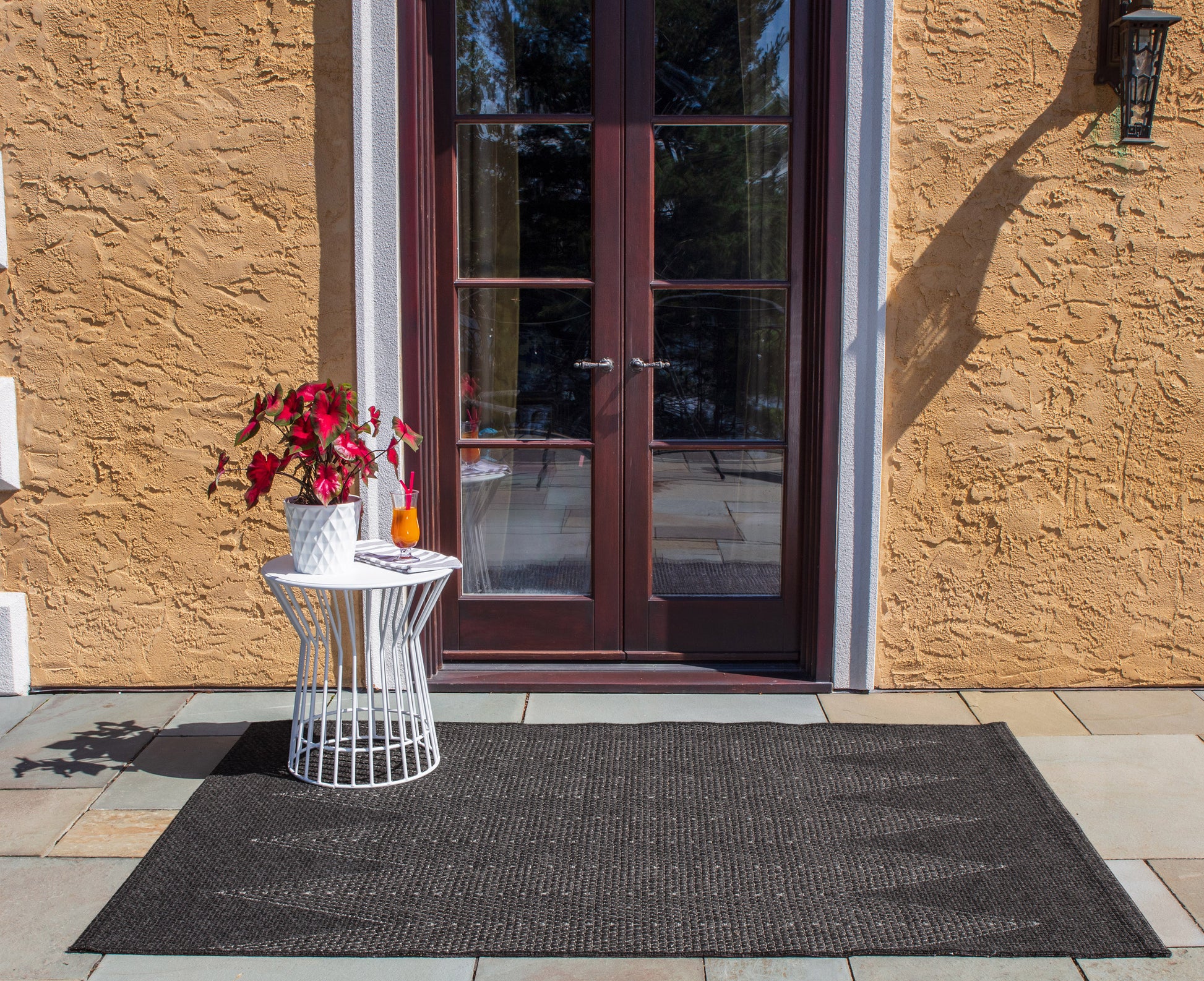 Brown French doors in a yellow stucco wall, white round metal table with potted plant & orange drink, black outdoor rug.