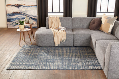 Blue-toned rug on the floor with a round wood side table holding a plant and a leaning abstract painting.
