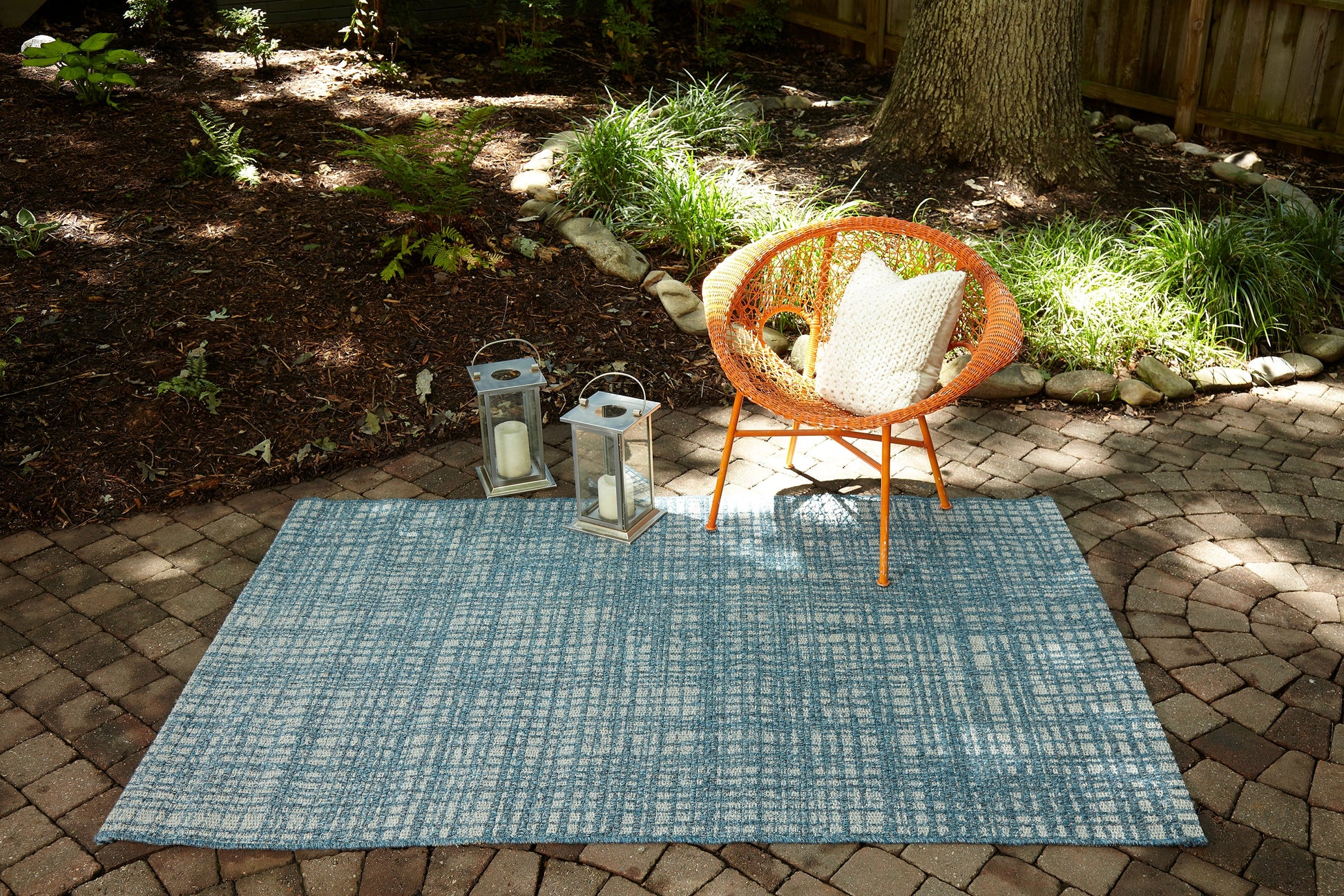 Blue patterned outdoor rug, 10' x 13', on a brick patio with an orange circular wicker chair and two metal lanterns in a garden setting.