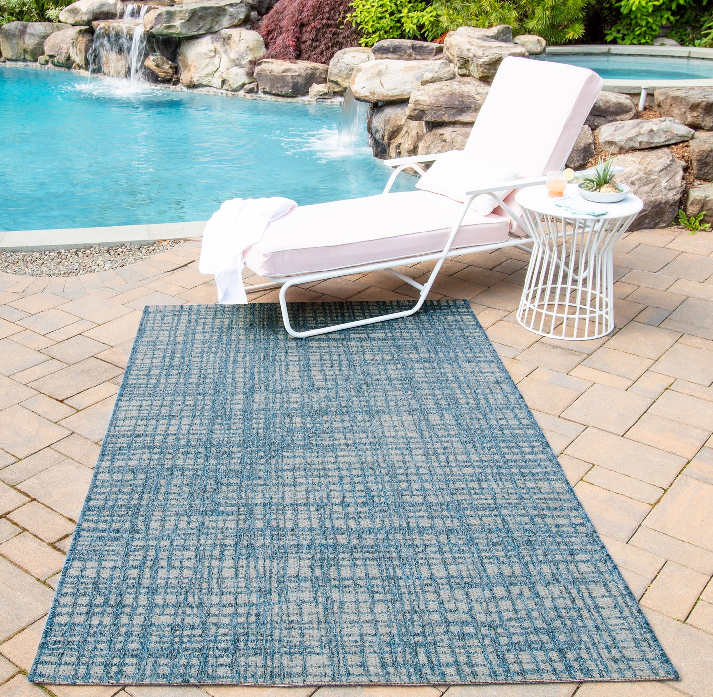 Blue patterned outdoor rug on stone patio beside white metal pool lounge chair with cushion.