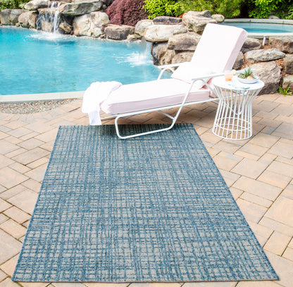 Rectangular blue outdoor rug with a subtle grid pattern, laid on tan pavers beside a poolside lounge chair.