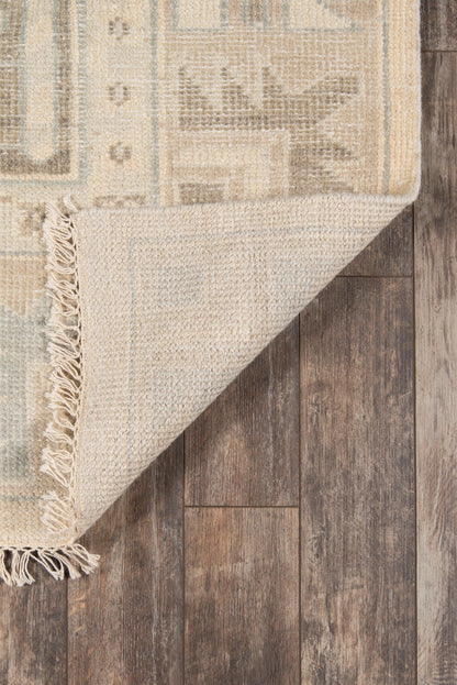 Beige woven area rug with light blue-gray geometric pattern and fringe, folded over dark wood floor.