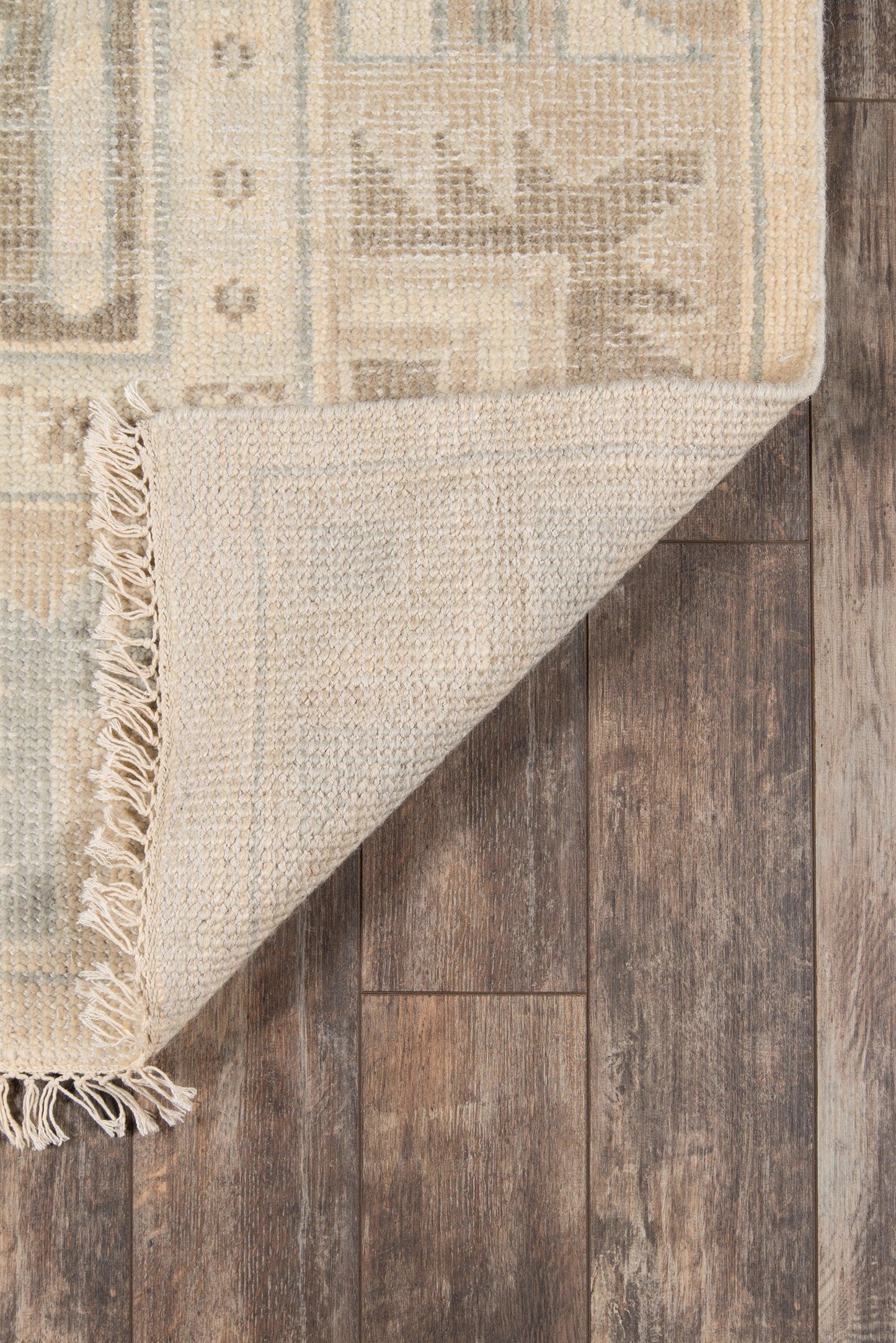Rectangular woven rug in cream, beige and pale blue with a geometric motif and fringe, partially folded on a dark wood floor.