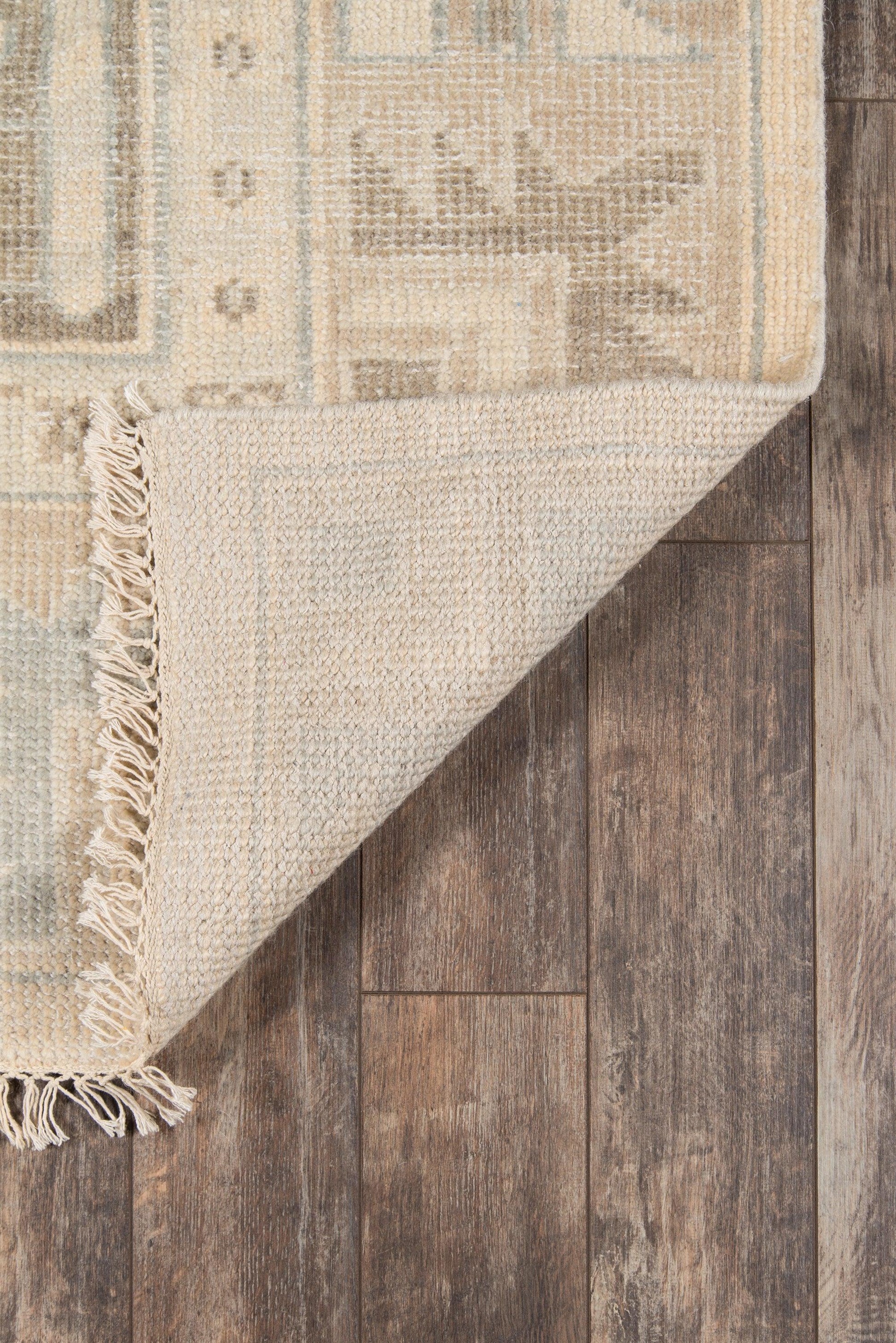 Beige and light blue patterned area rug with fringe edge, folded corner on dark wood floor.