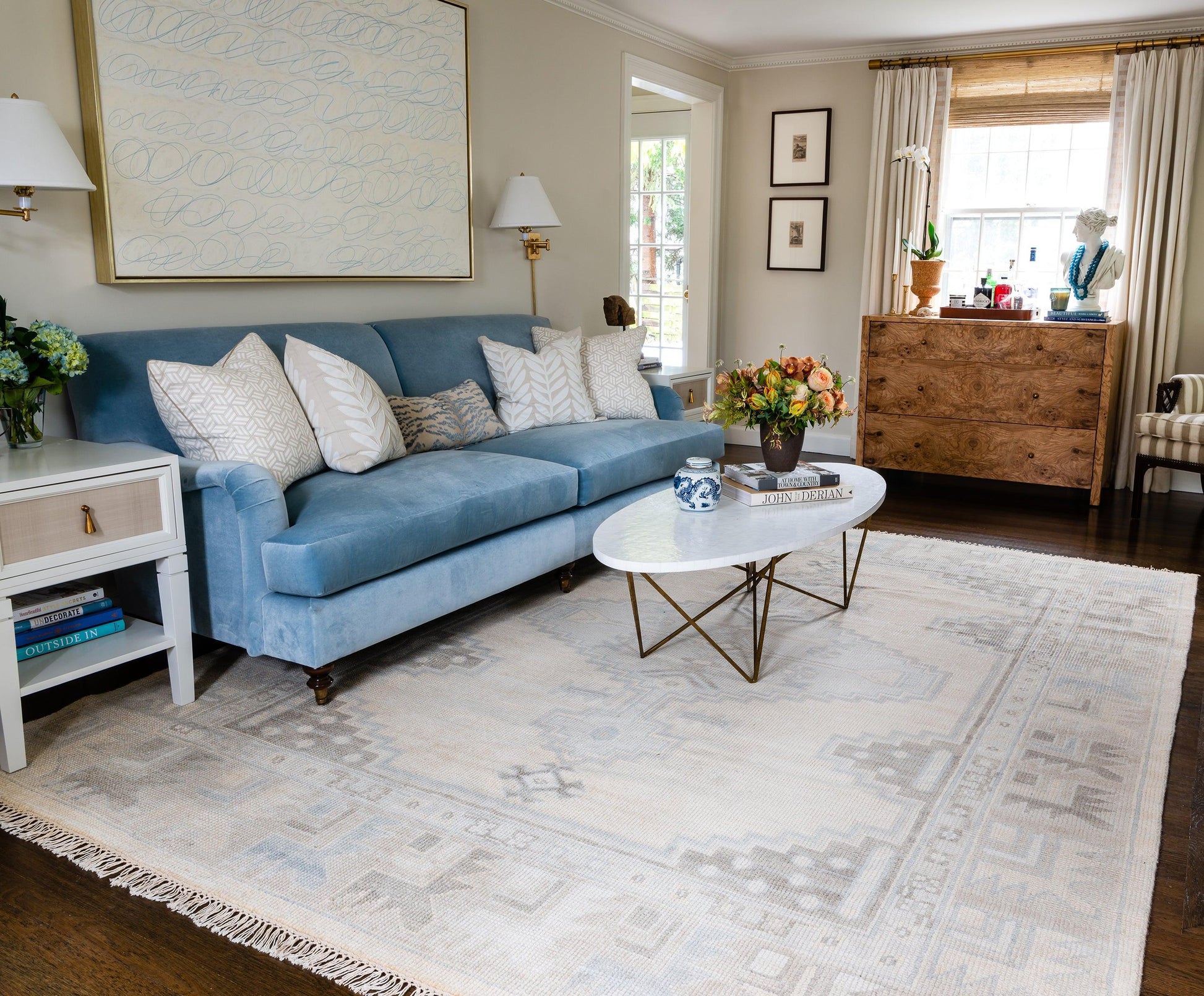 Light blue sofa with white pillows, white round coffee table with gold legs on a beige geometric rug in a living room.
