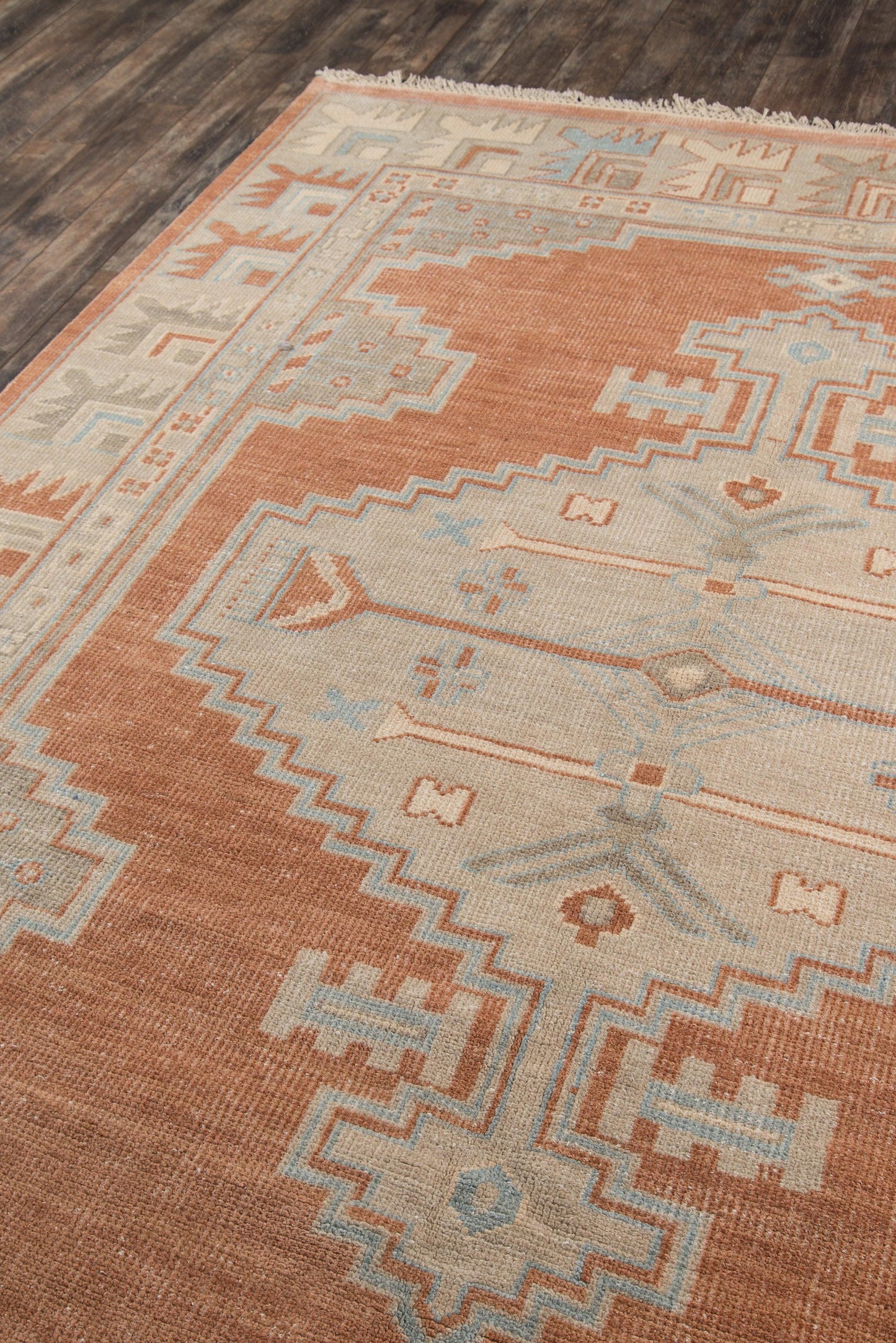 Rust and cream kilim-style wool rug with light blue geometric motifs and zigzag border, fringed edges on dark wood floor.