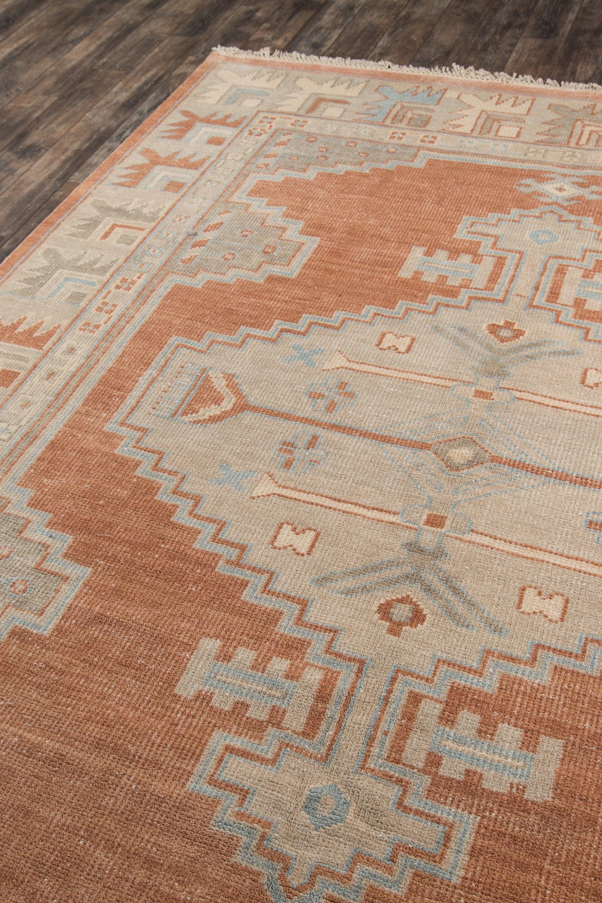 Rust and cream kilim-style wool rug with light blue geometric motifs and zigzag border, fringed edges on dark wood floor.