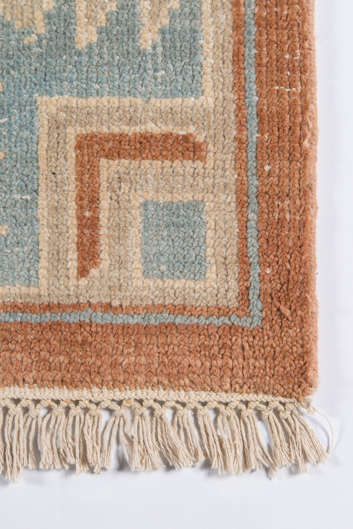 Close-up of a woven rug corner with a geometric border in blue, beige and rust, cream fringe.