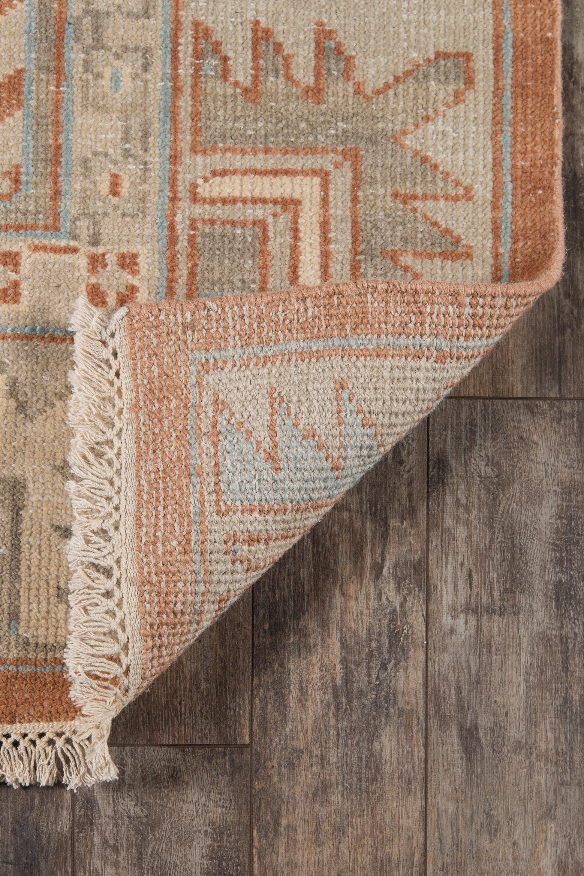 Boho geometric woven area rug in warm beige, rust & pale blue with fringe on a wood floor.