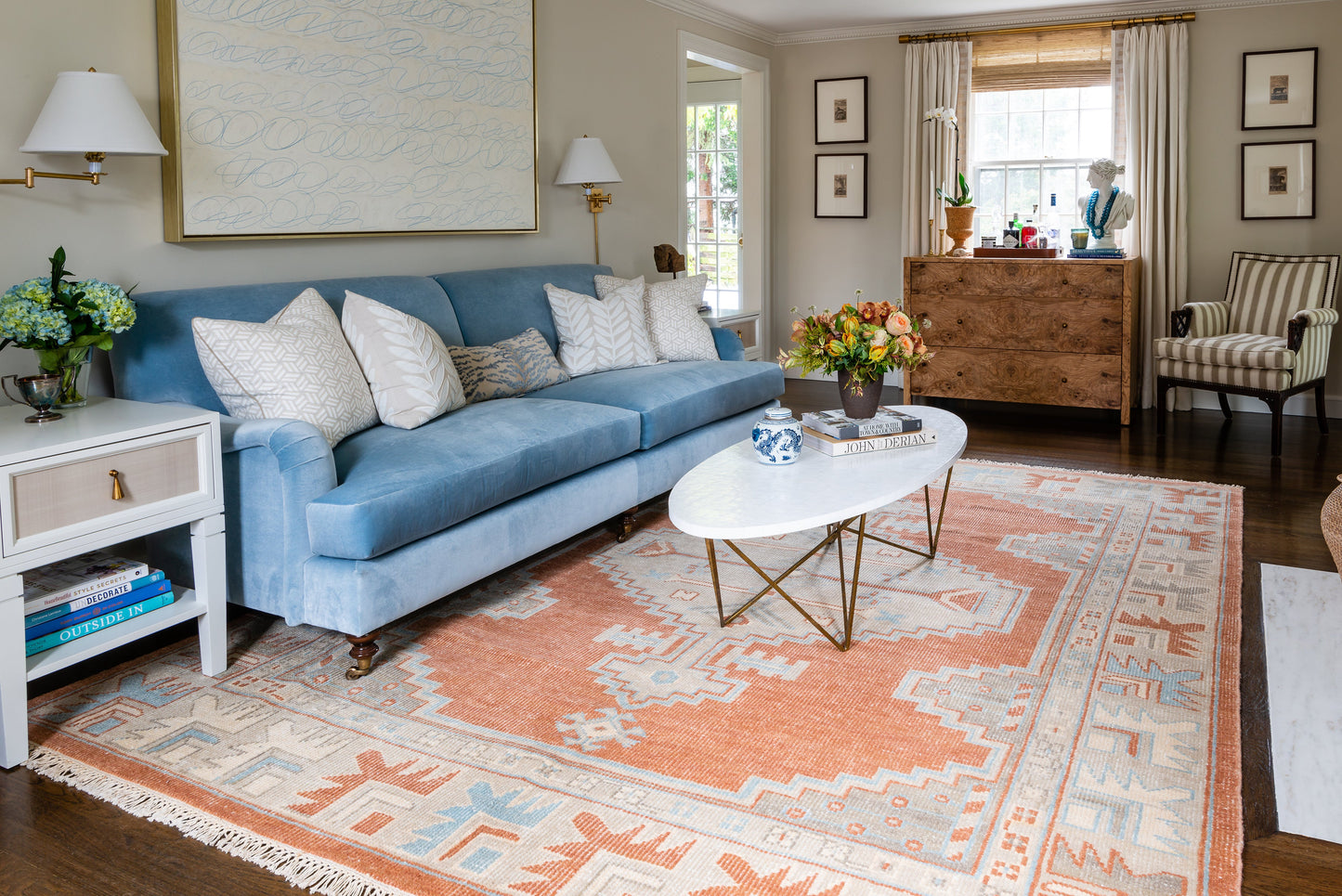 Cozy living room with a blue velvet sofa, white patterned cushions, oval marble coffee table, peach geometric rug and brass lamps.