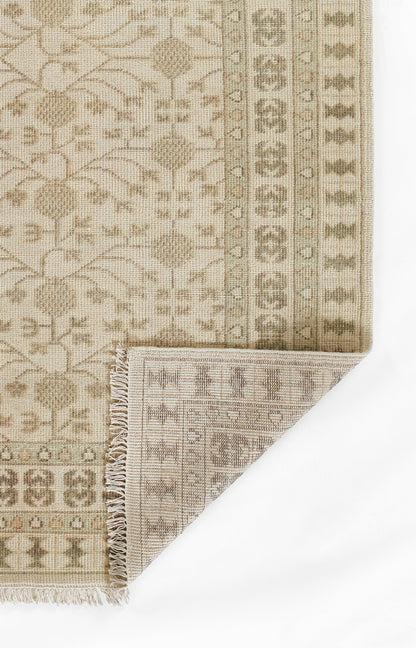 Beige oriental rug with floral vines & geometric borders; folded corner reveals fringe trim.