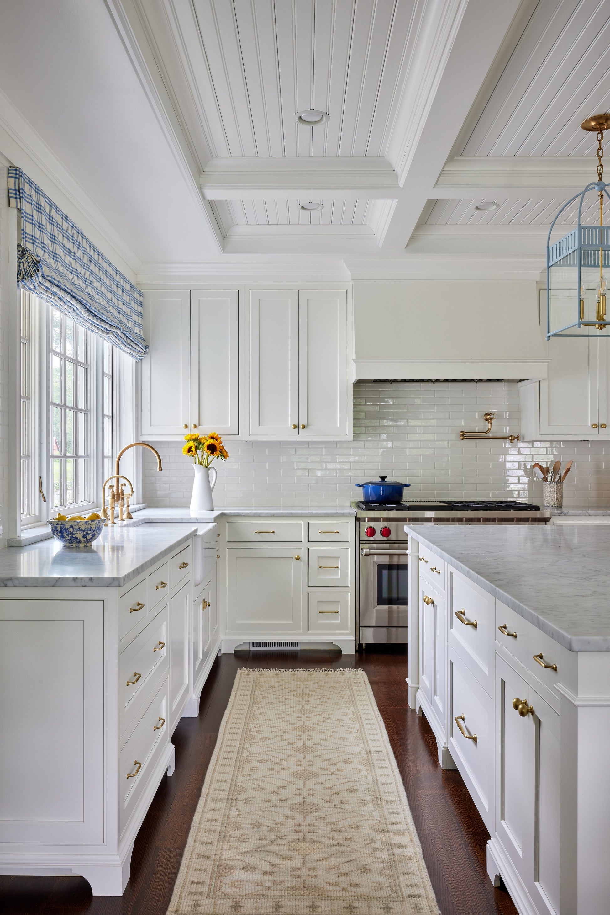 Bright white kitchen with coffered ceiling, marble countertops, brass hardware, window valance, sunflowers, and island.