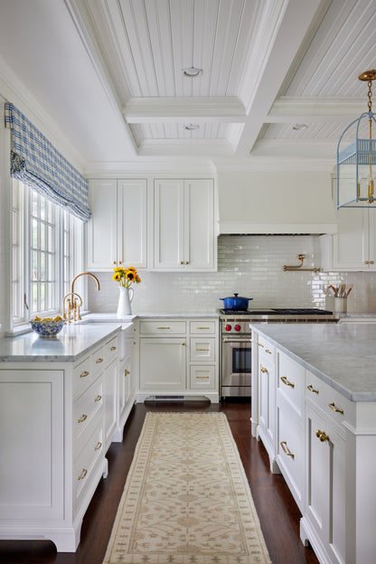 Bright white kitchen with coffered ceiling, marble countertops, brass hardware, window valance, sunflowers, and island.