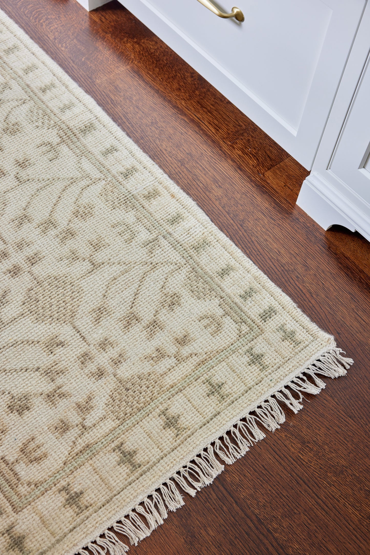 Beige traditional patterned area rug with cream fringe on dark hardwood floor beside white cabinetry.