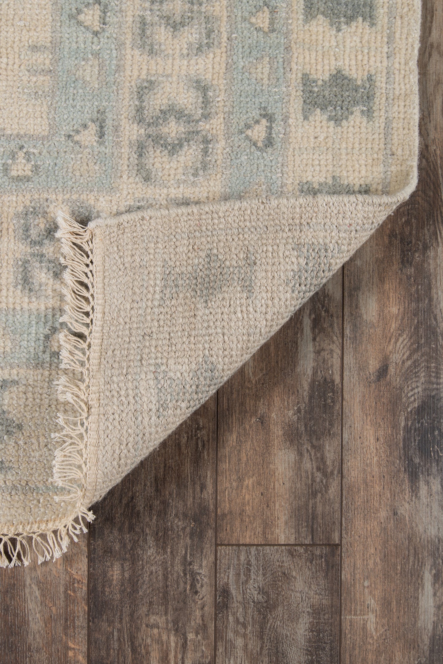 Ivory and muted teal-blue woven area rug with geometric pattern and fringe, folded corner on wood floor.