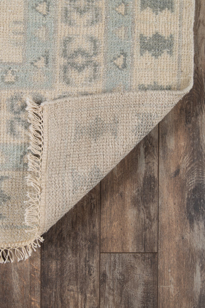Ivory and muted teal-blue woven area rug with geometric pattern and fringe, folded corner on wood floor.