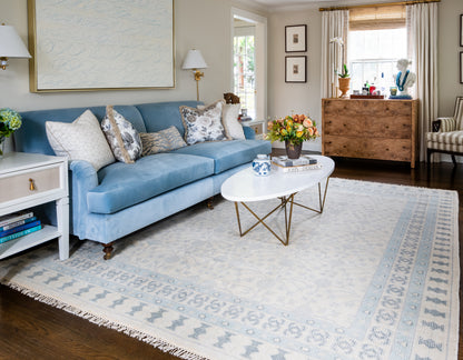 Blue velvet sofa with patterned cushions, oval white marble coffee table with gold legs, blue and cream geometric rug in living room.