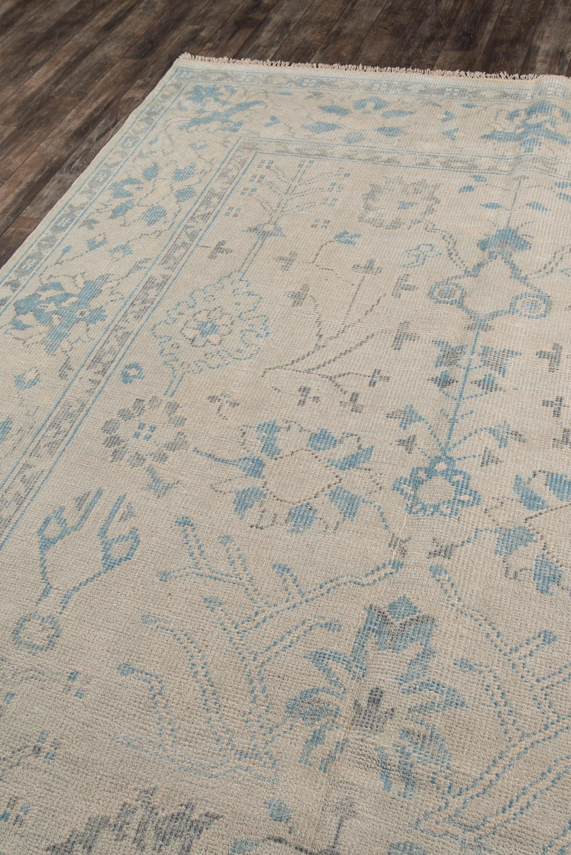 Beige oriental rug with pale blue floral motifs & border, fringed edge on dark wood floor.