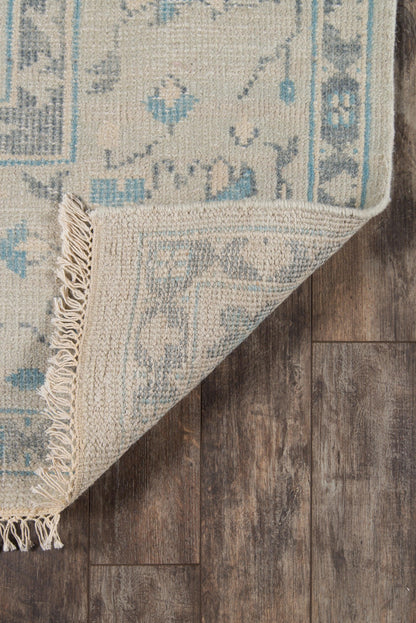 Beige oriental-style rug with light blue ivy motifs and fringe, folded over on dark wood floor.