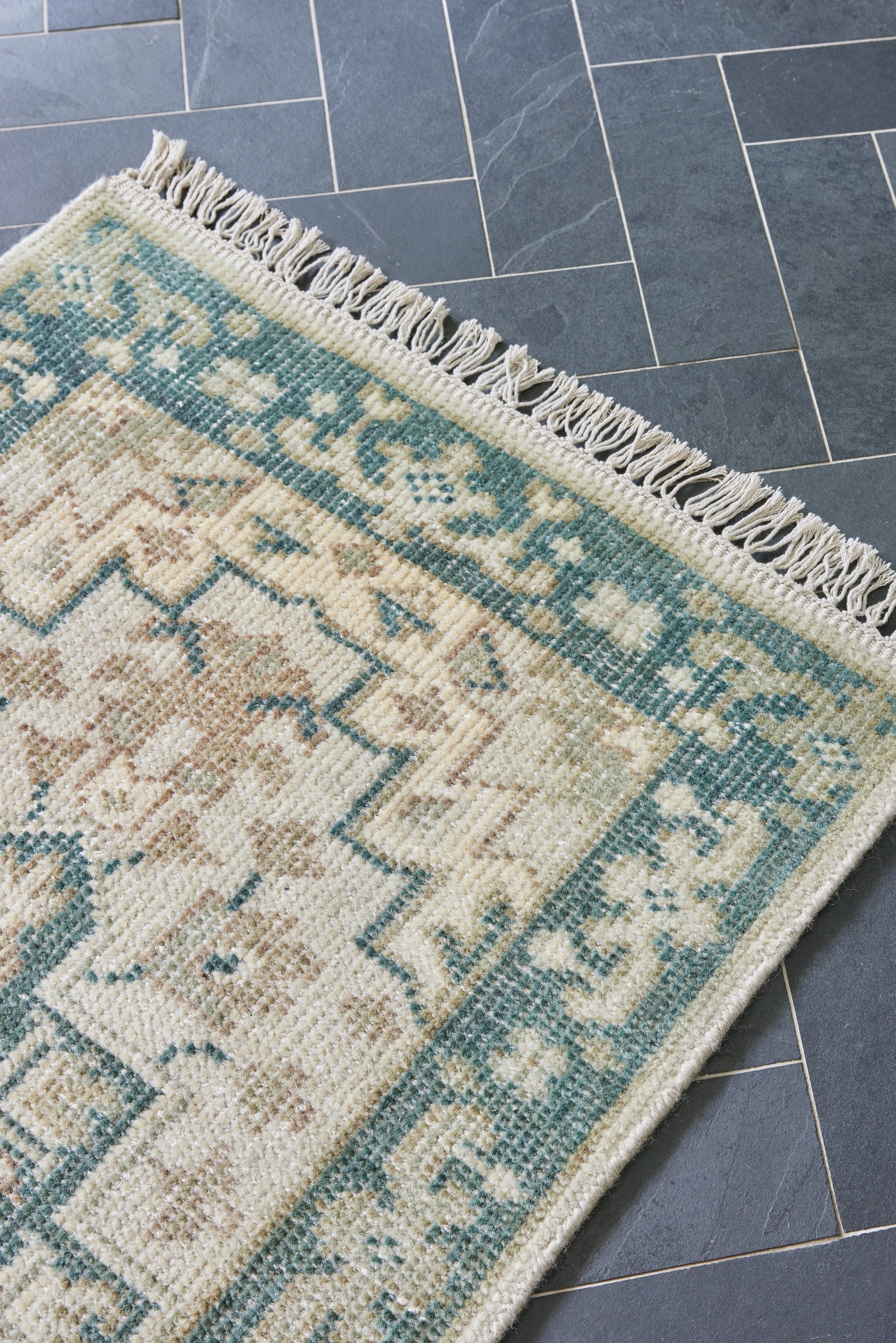 Cream and teal oriental rug with geometric floral pattern and fringed edge resting on blue-gray tiled floor.