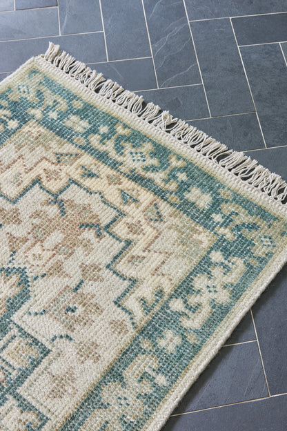 Cream and teal oriental rug with geometric floral pattern and fringed edge resting on blue-gray tiled floor.