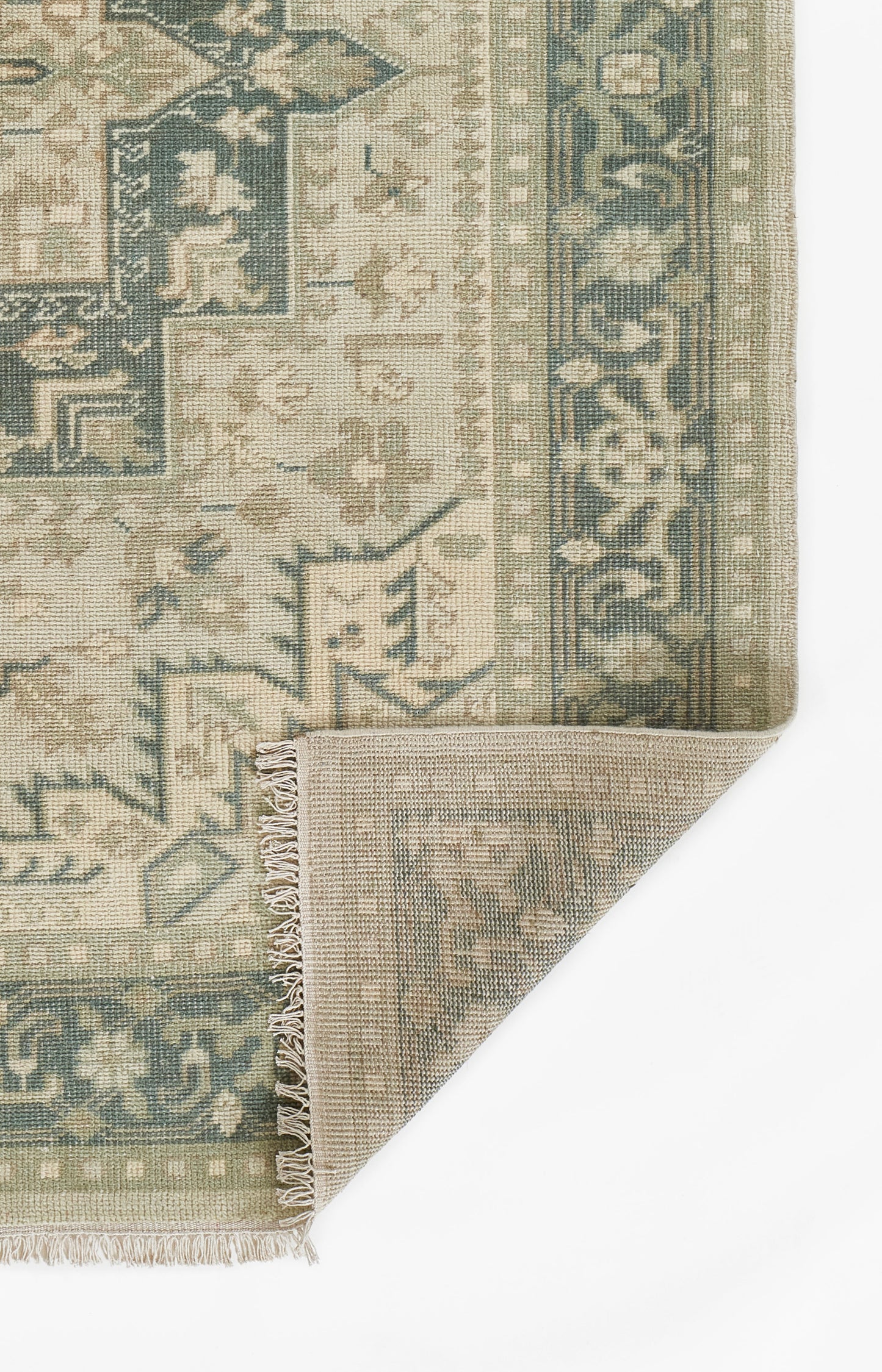 Persian-inspired woven area rug in green, blue, and beige with ornate border and fringe, folded corner.