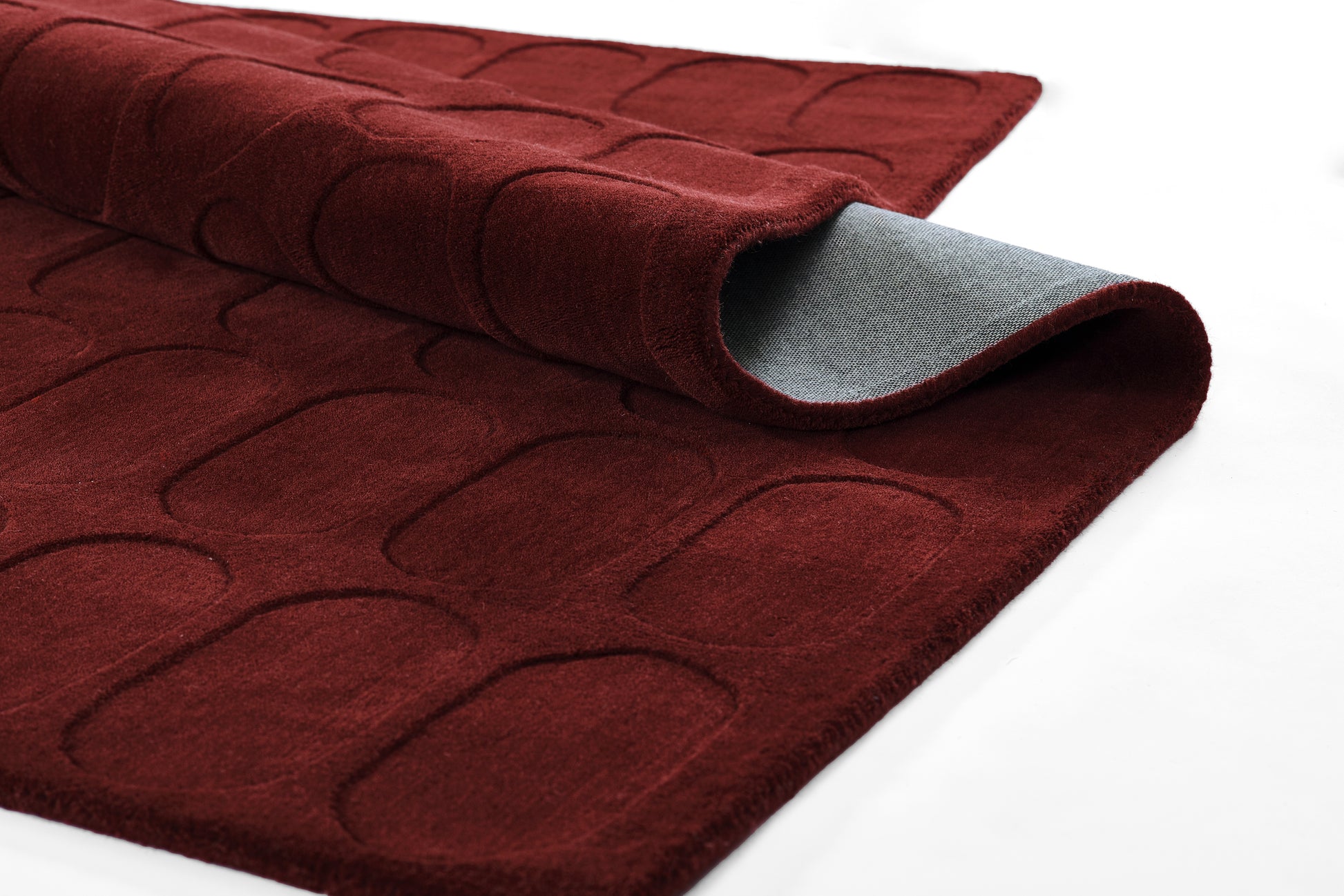 Deep red tufted area rug with curved rectangular quilting and rolled edges; gray underside visible; plush decorative floor mat.