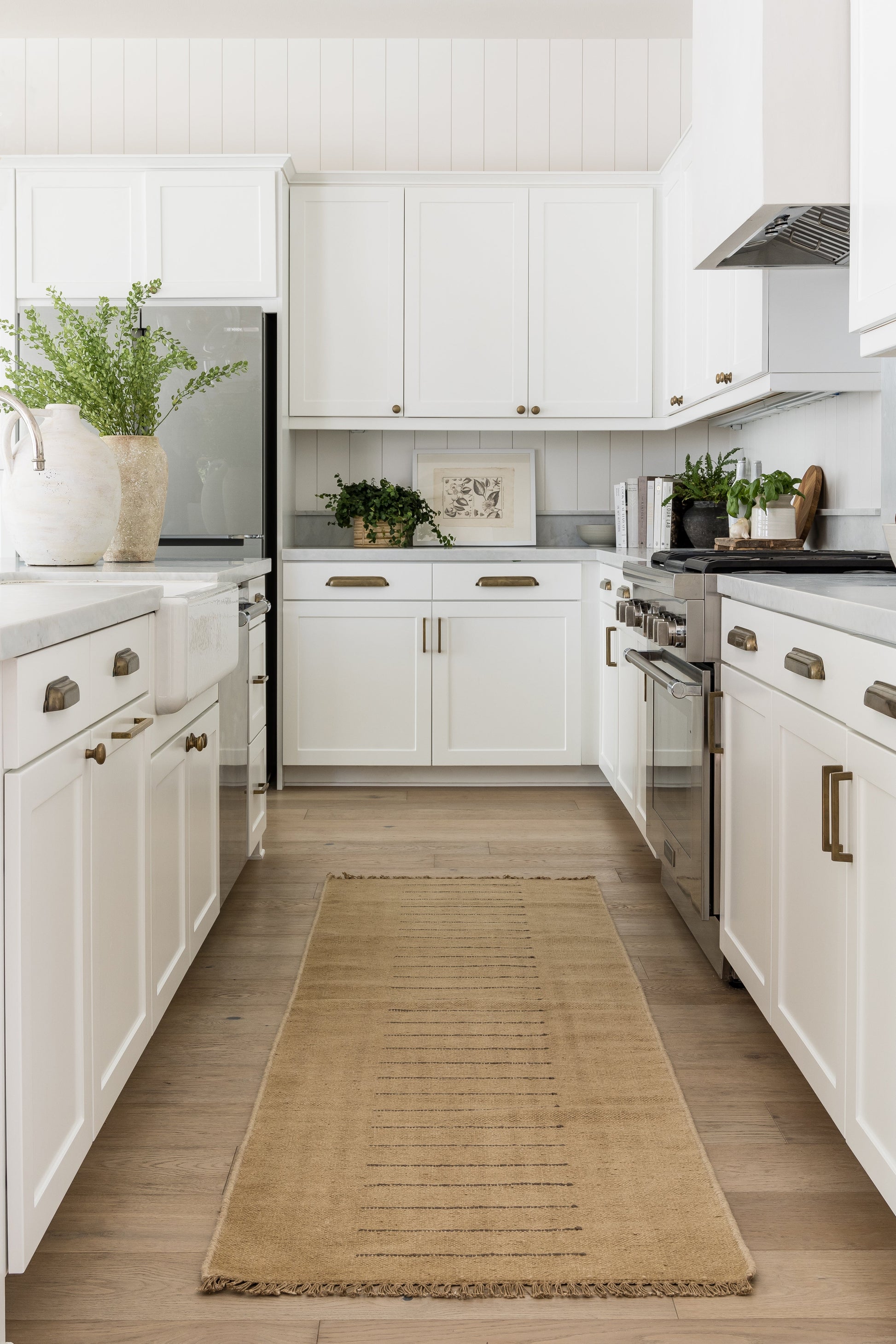 Bright white kitchen with white cabinets, stainless steel appliances, light wood floor, beige runner rug, and greenery.