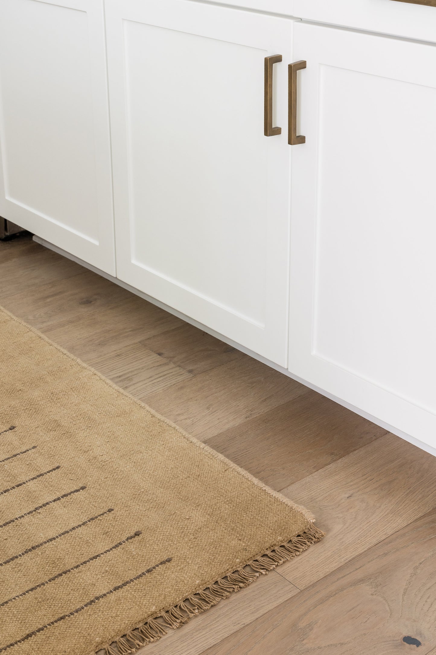 White shaker cabinets with brass bar pulls, light wood flooring, and a beige fringed area rug.