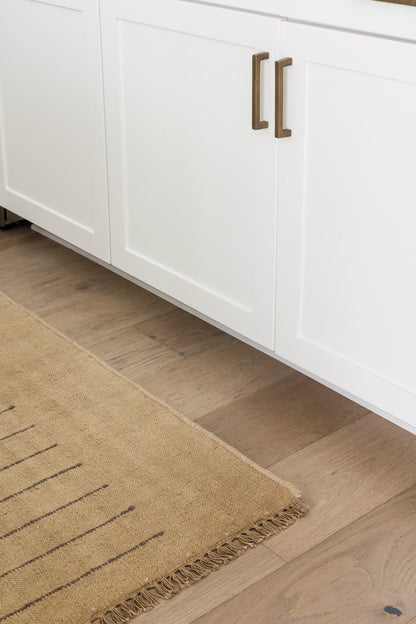 White shaker cabinets with brass bar pulls, light wood flooring, and a beige fringed area rug.
