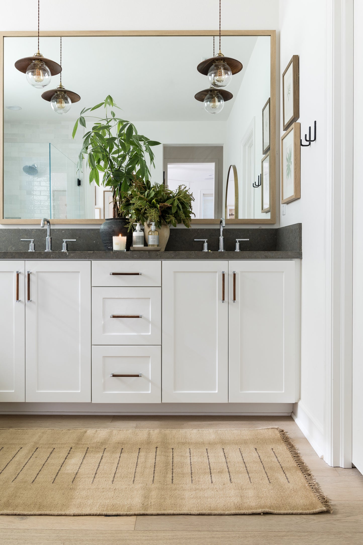 White shaker vanity with dark countertop, copper handles, large wood-framed mirror, hanging globe lights, and leafy plants.