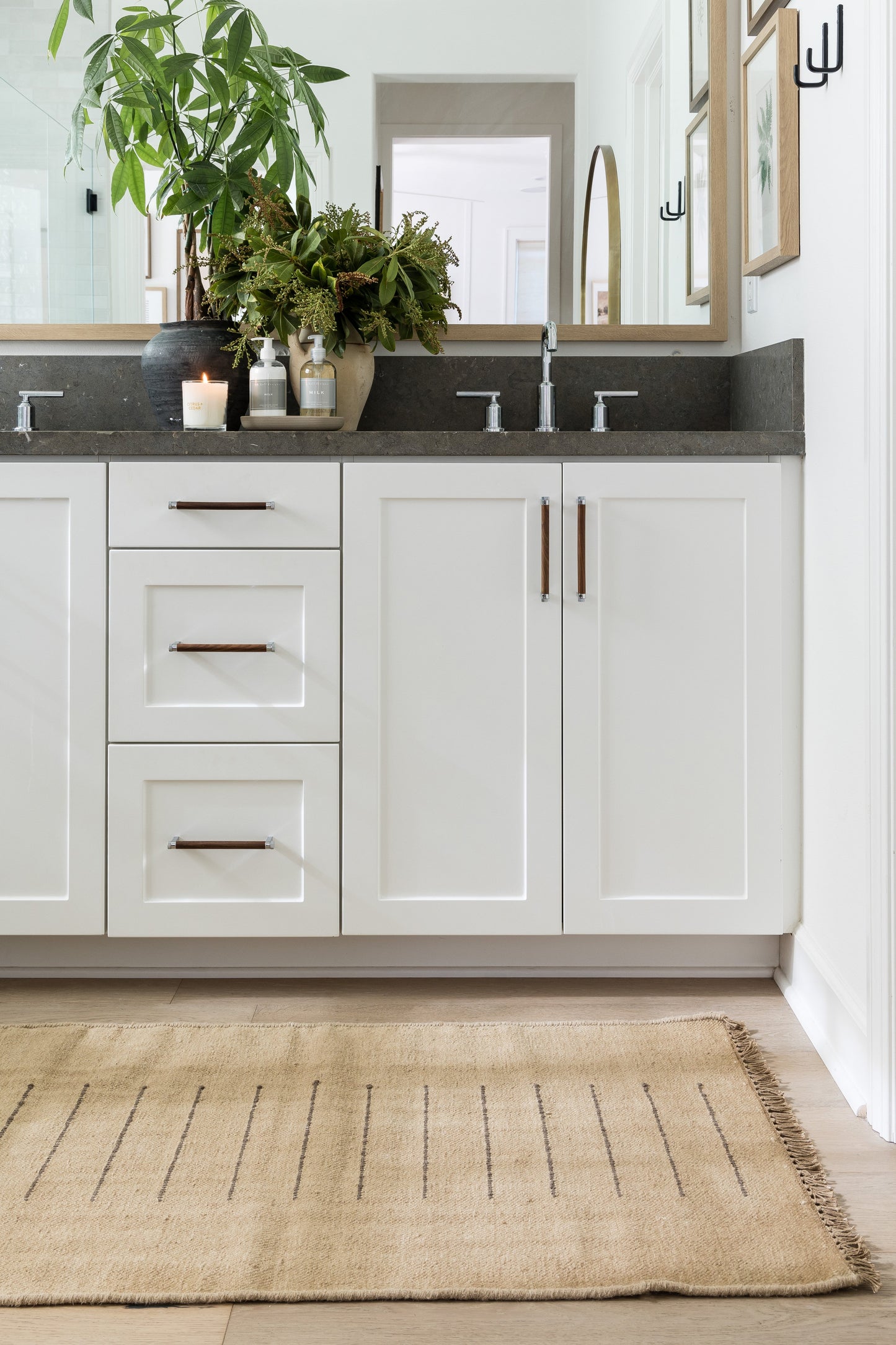 White shaker vanity with three drawers left, double doors right, gray countertop, two faucets, copper handles, plants, rug.