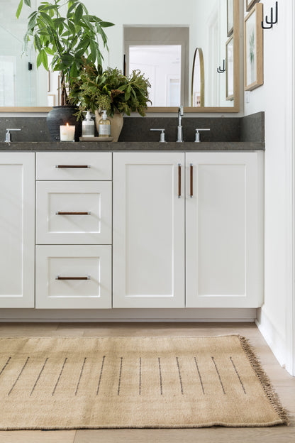 White shaker vanity with three drawers left, double doors right, gray countertop, two faucets, copper handles, plants, rug.