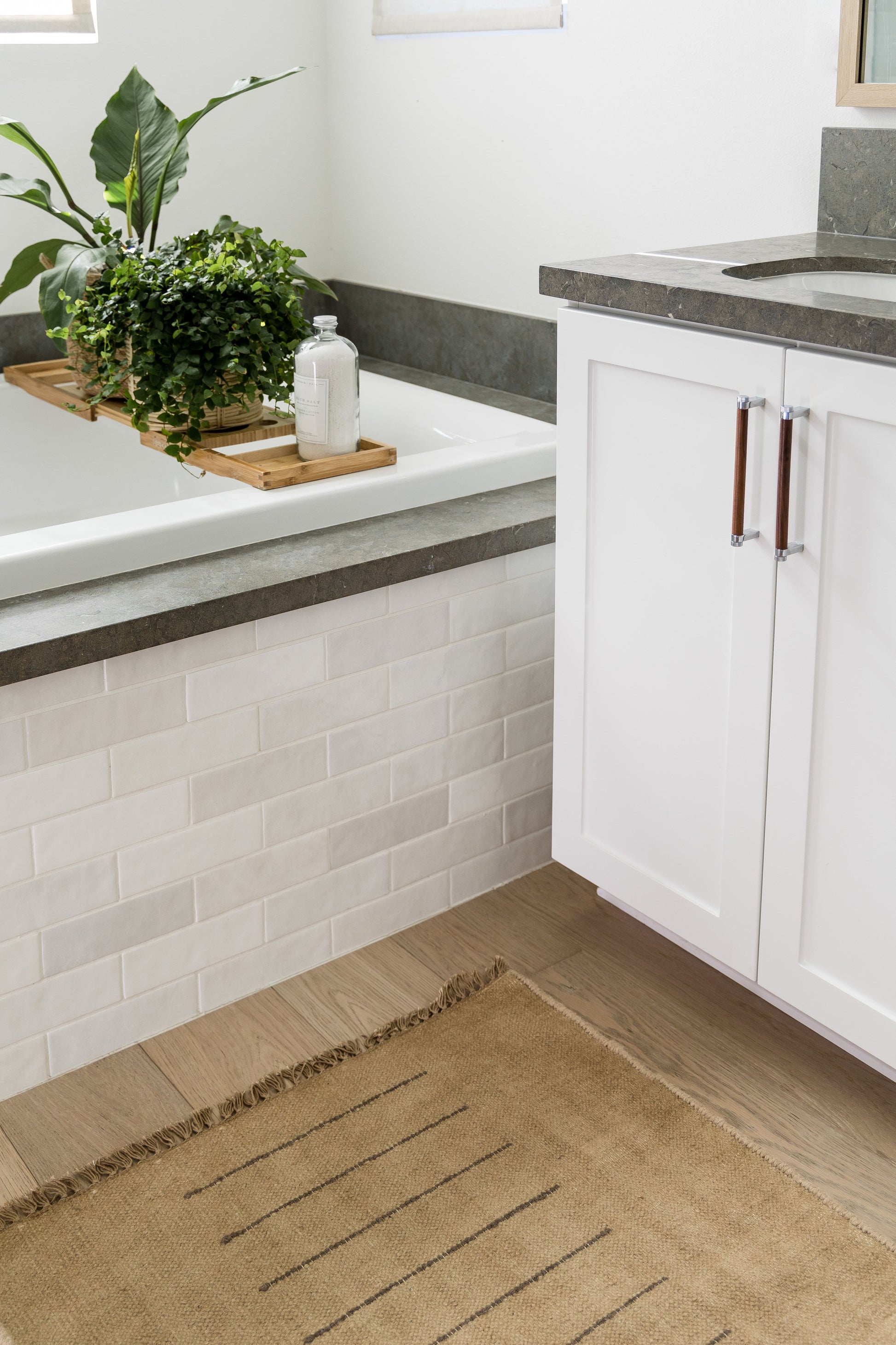 White bathroom vanity with dark stone countertop, tub with wooden tray, potted plants, bottle, white subway tiles, beige rug.
