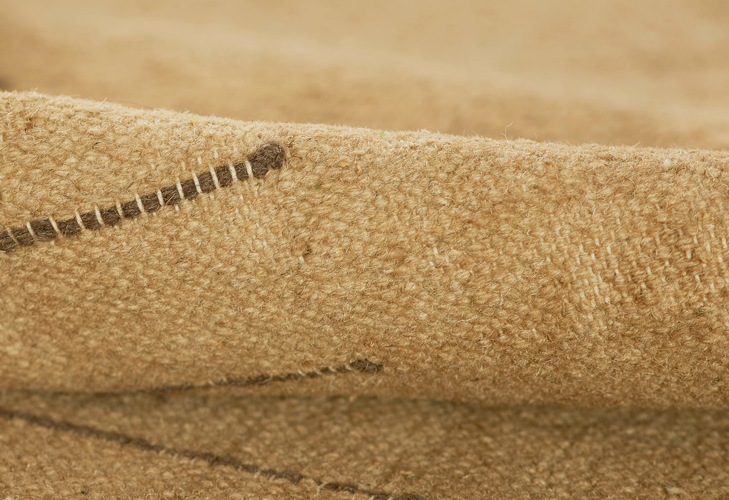 Close-up of beige burlap-like rug texture with visible stitching and rough woven jute fibers.
