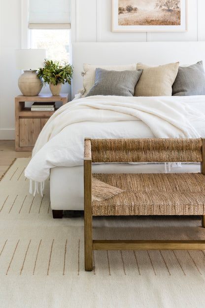 White bed with cream throw and neutral pillows, woven rattan bench at foot, wooden nightstand with lamp and plant.
