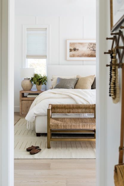 Ivory woven rattan bench with wooden legs at the foot of a white, cozy bed in a neutral bedroom.