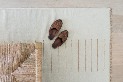 Light beige rug with brown vertical stitched lines and woven fringe edge; pair of dark brown woven slippers on it.