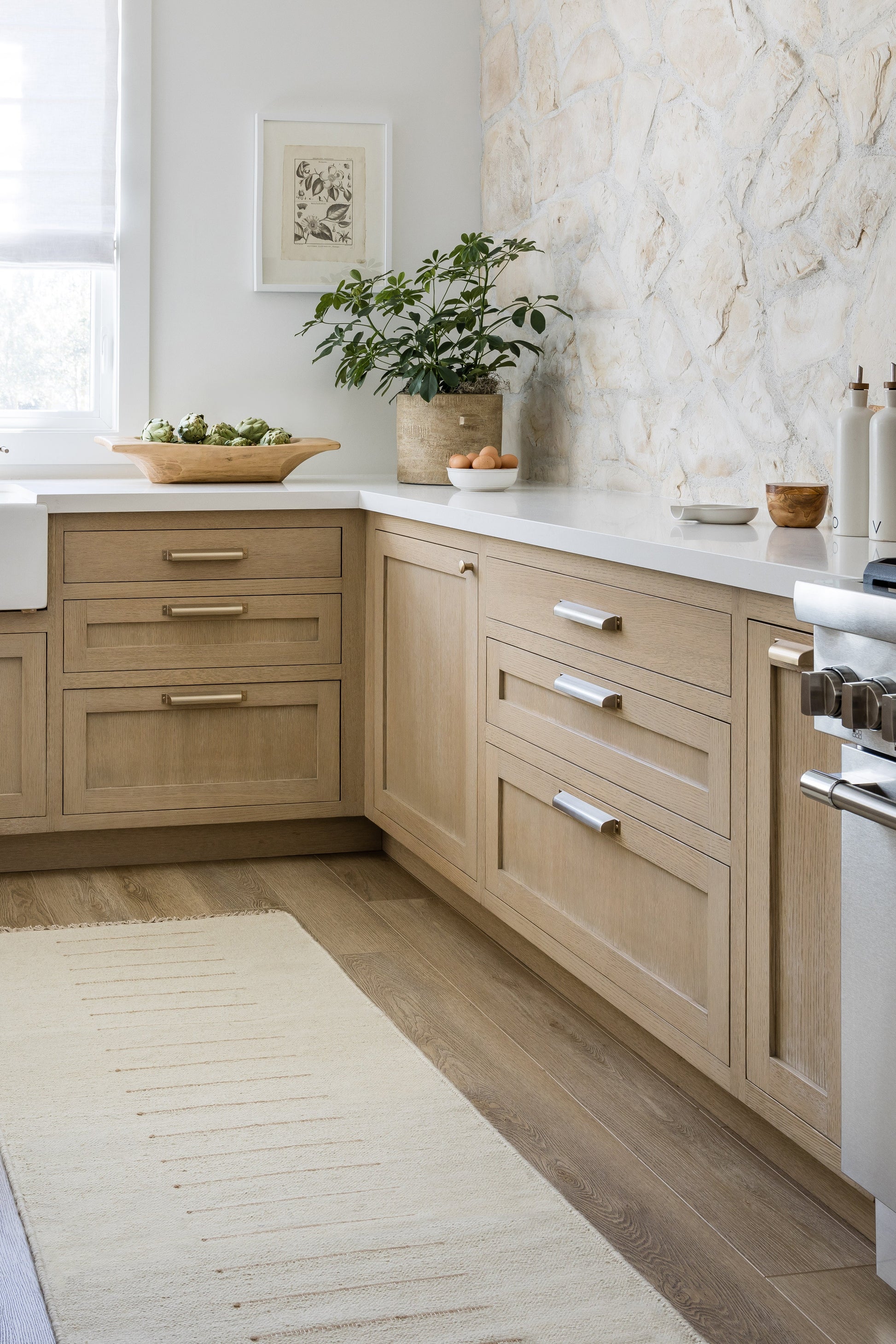 Beige wood kitchen cabinets with white countertops, potted plant, stone-textured wall, wooden floor, and a stainless stove.