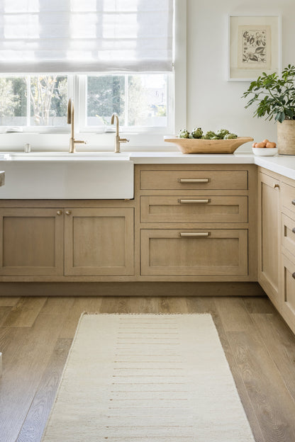 Light beige rug on a light wood floor with a few green plants nearby.