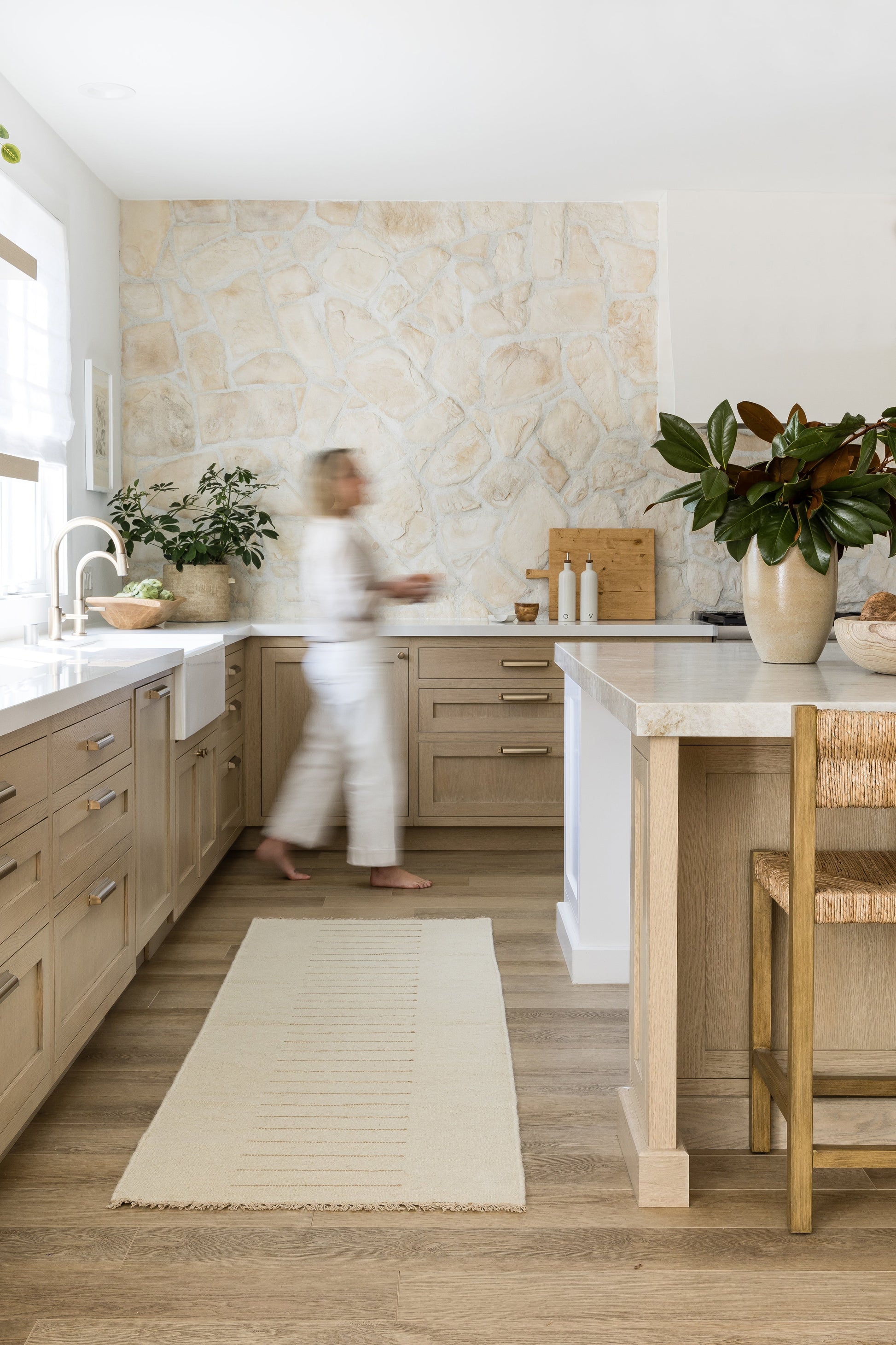 Bright modern kitchen with light wood cabinets, beige stone wall, island, stone countertop, potted plants, and wooden stools.