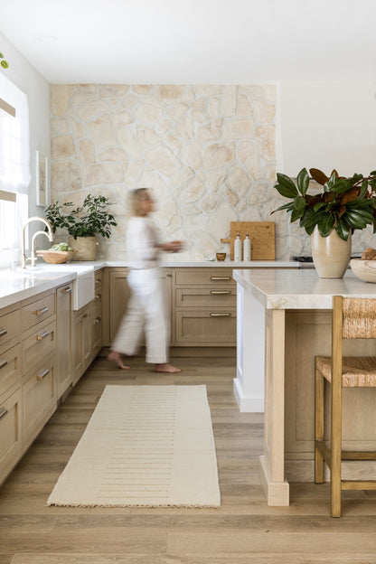 Bright modern kitchen with light wood cabinets, beige stone wall, island, stone countertop, potted plants, and wooden stools.